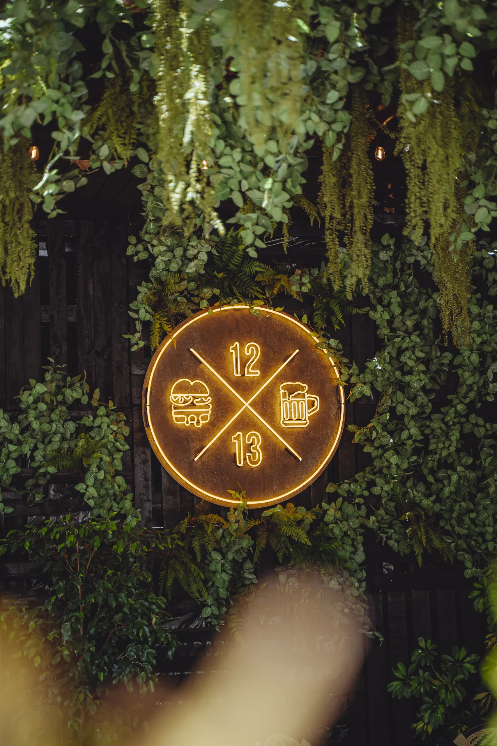 Letrero luminoso de Sanguchería DoceTrece en Chile con logo 12 13, ícono de hamburguesa y cerveza en muro verde con plantas y ambiente nocturno