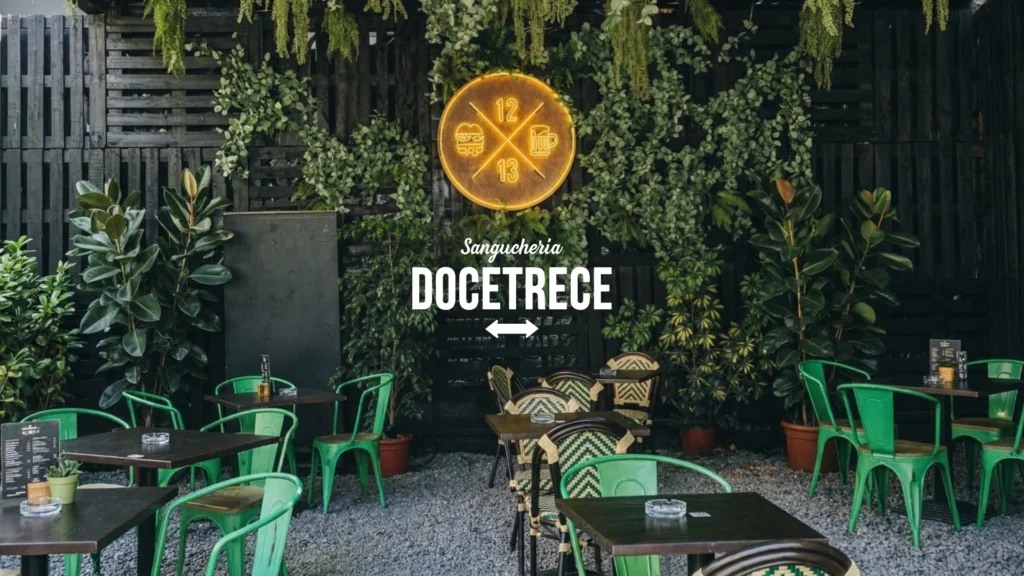 Terraza al aire libre de restobar en santiago Sanguchería DoceTrece en Santiago de Chile, mesas y sillas verdes, muro vegetal y letrero luminoso 12 13 en el fondo